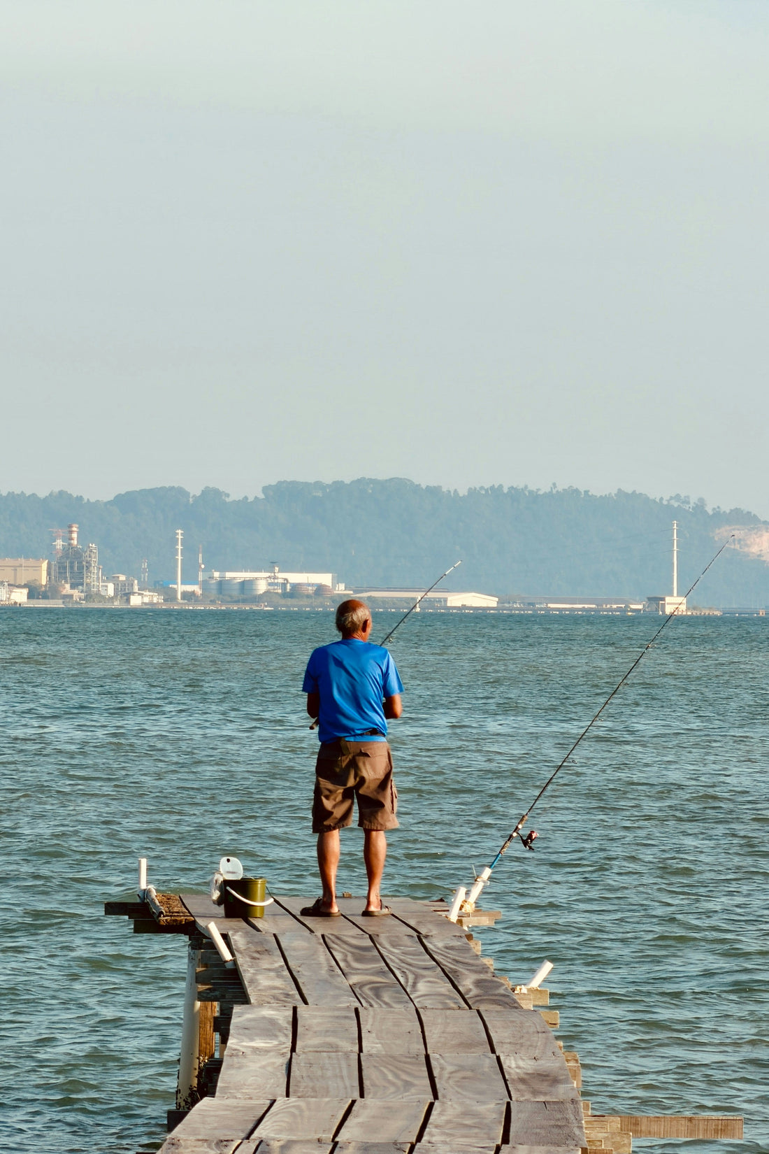 Man fishing off of pier with factory across the harbor