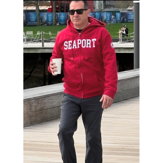 Man walking with coffee on marina wearing a red Seaport zip hoodie
