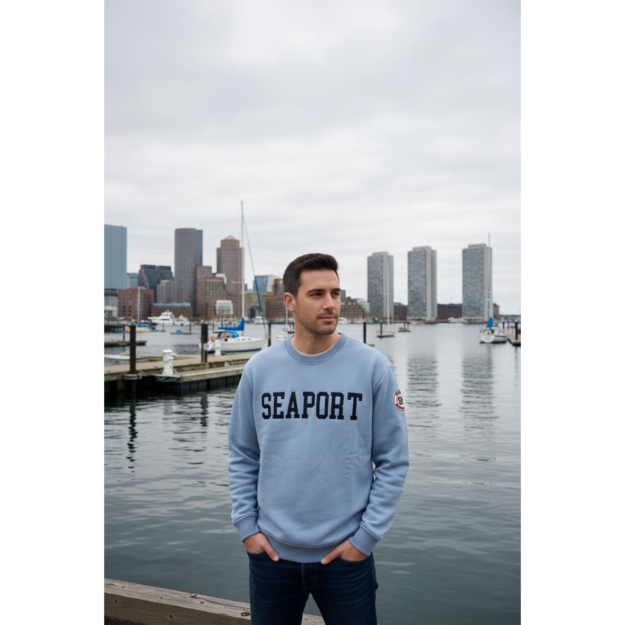 A light blue crewneck sweatshirt with 'SEAPORT' printed in black across the chest and a circular 'SEAPORT' patch on the right sleeve, hanging on a white hanger against a teal corrugated metal background.