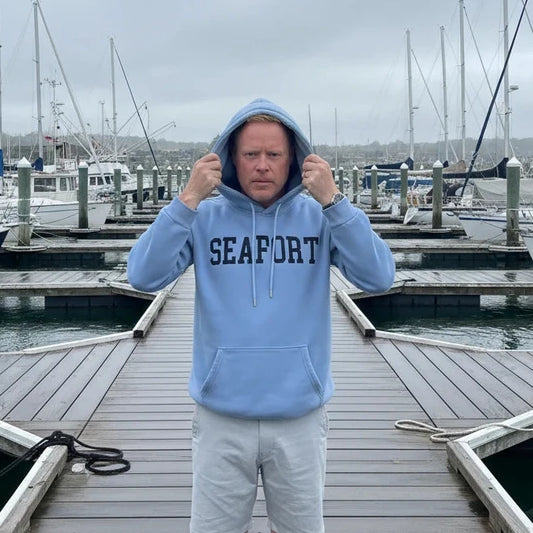 Person wearing a light blue heavy hoodie with 'SEAPORT' text, standing on a waterfront marina