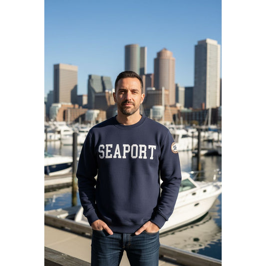 Man wearing a navy sweatshirt with 'SEAPORT' text, standing in front of a city skyline with boats.